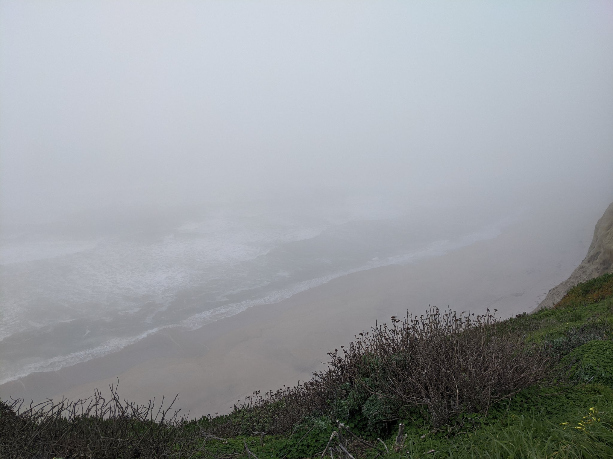 A photo of a beach taken from the top of a clif. The image is shrouded in fog, but some small waves can be seen.