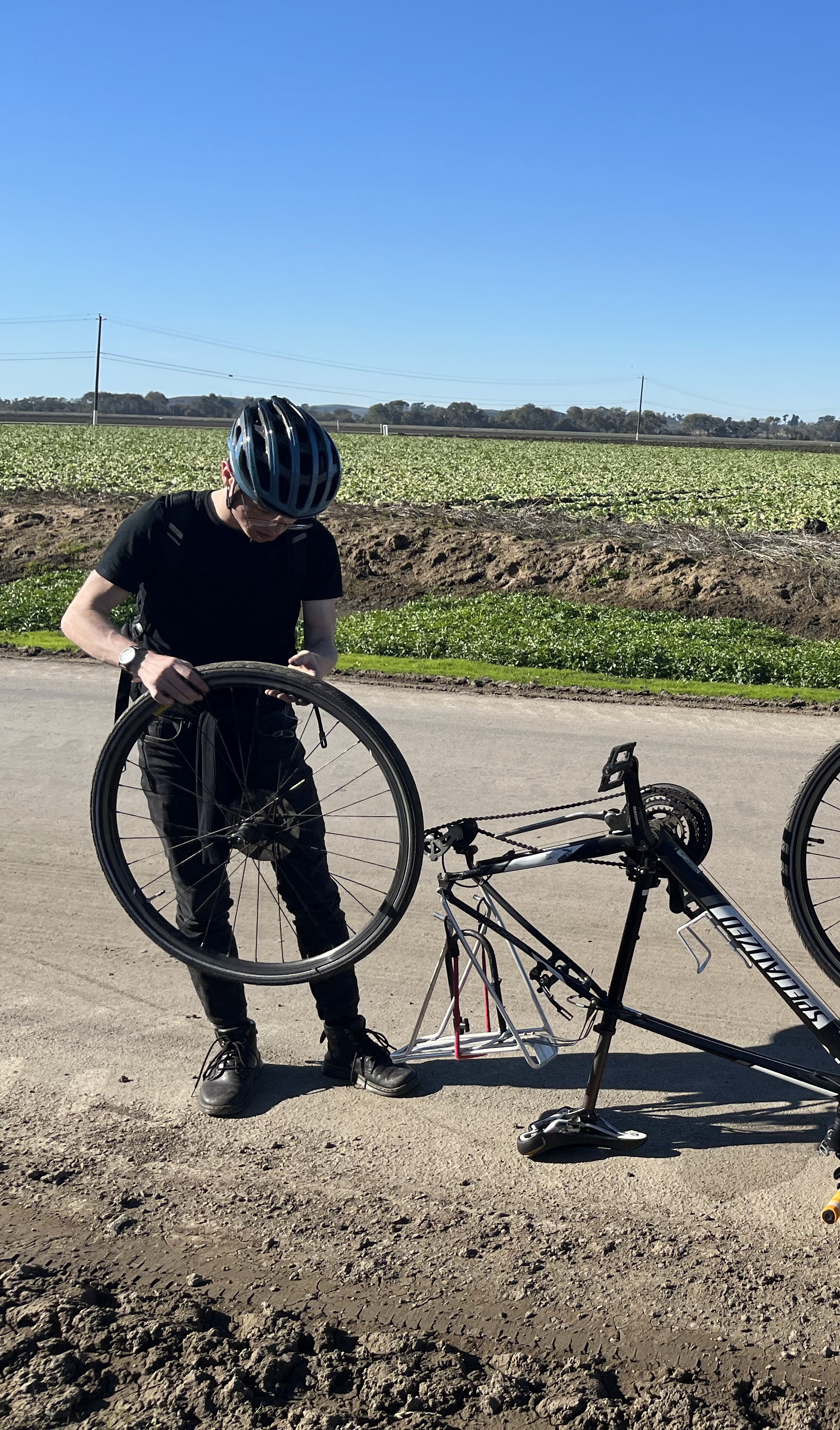 A photo of Wesley Aptekar-Cassels standing on a dirt road fixing a flat tire on a bike.