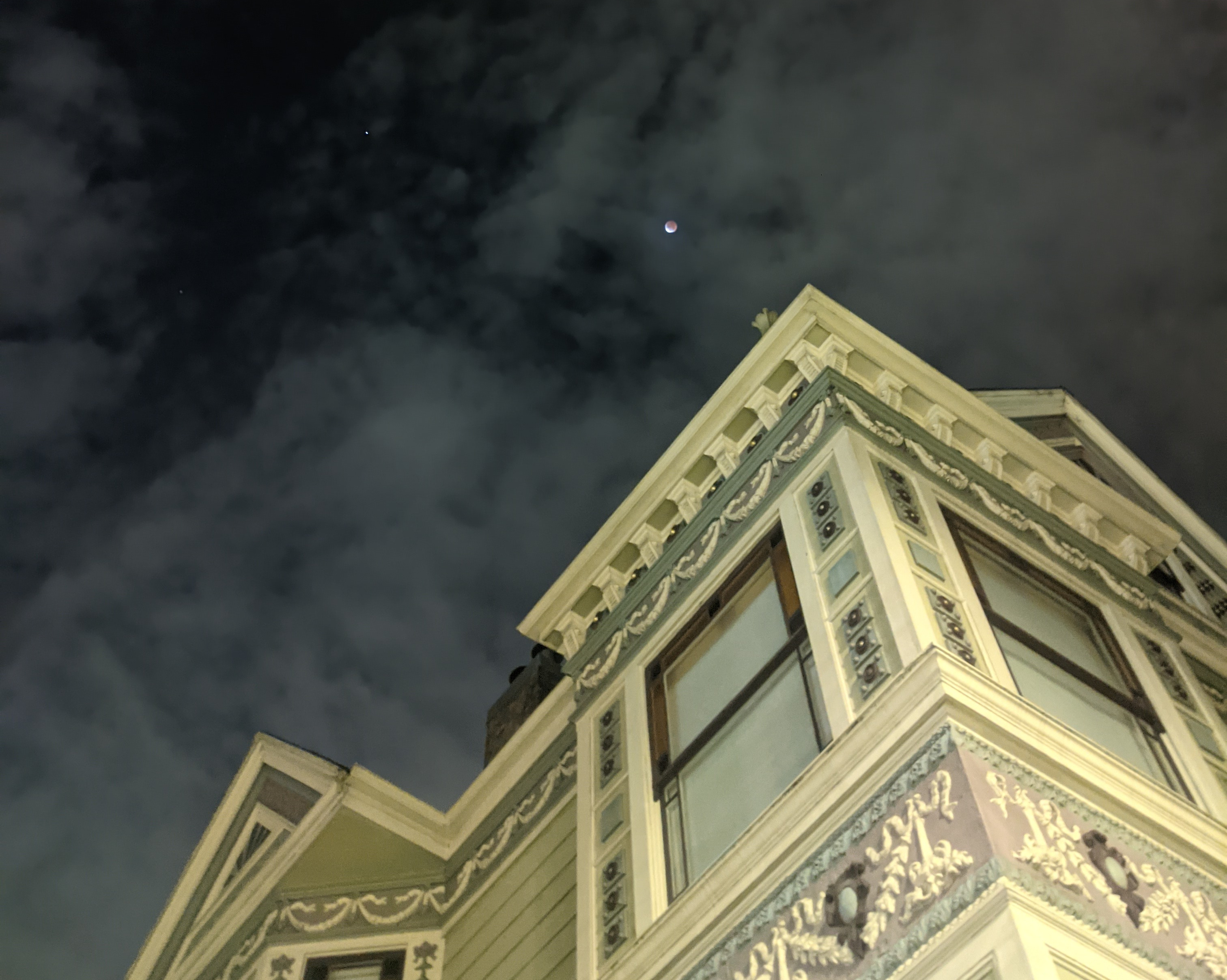 A very small and fuzzy picture of a lunar eclipse next to a green victorian house.
