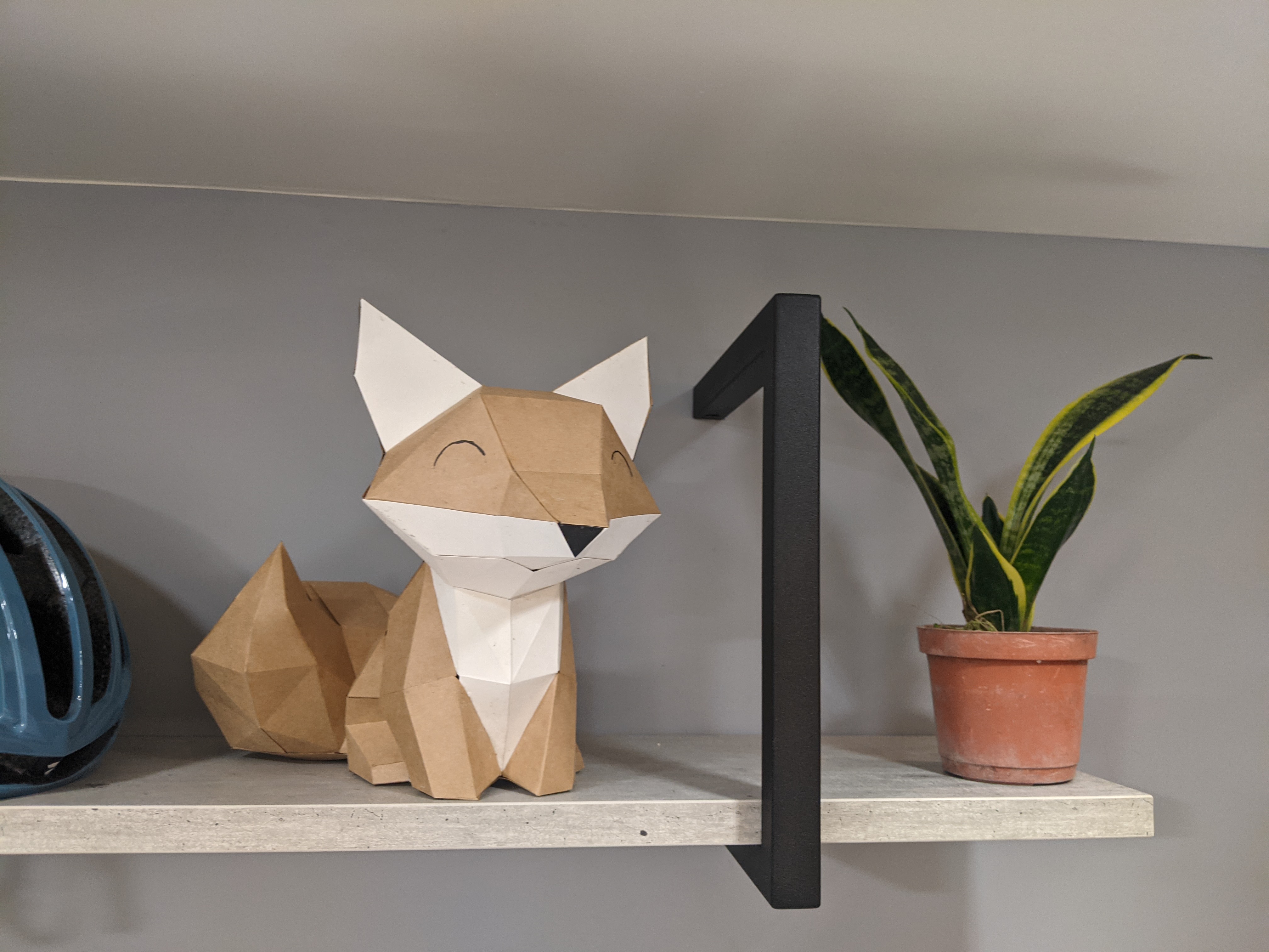 A cute papercraft fox, sitting on a shelf next to a plant and a bike helmet.