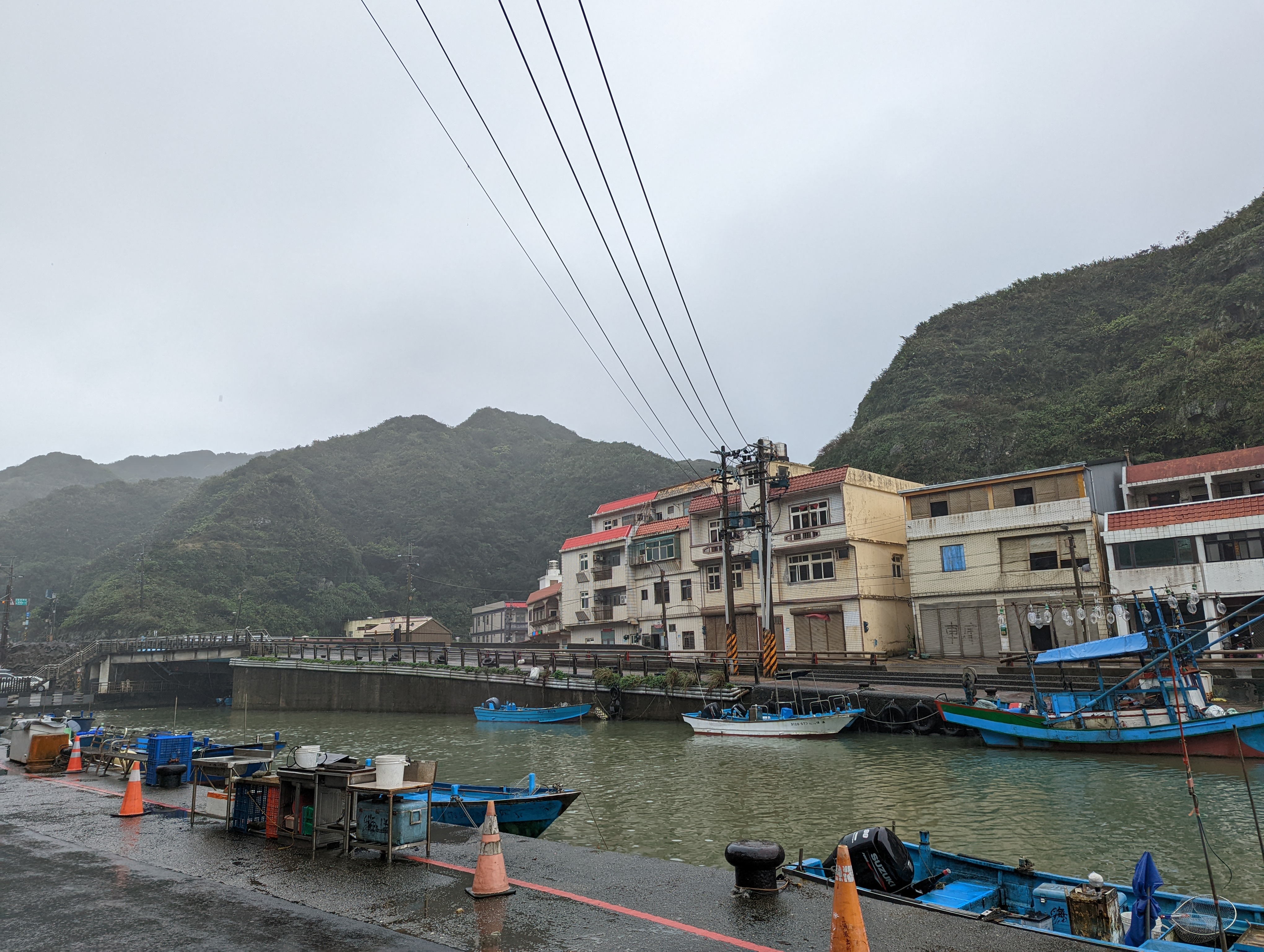 A photo of a small canal with some boats docked. Across the shore are some buildings, and mountains behind them. It's foggy, and some power lines stretch across the canal.