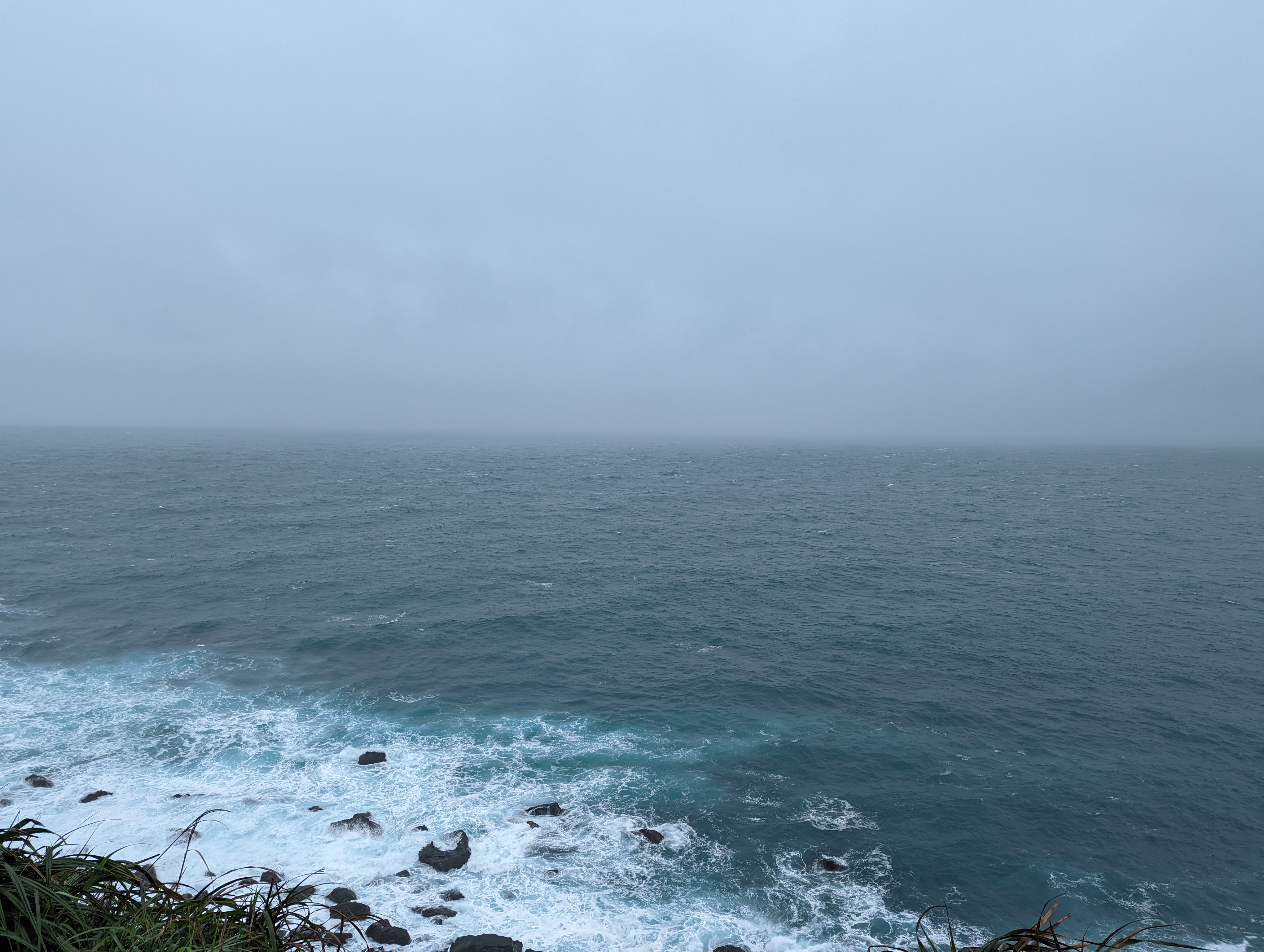 A photo of the ocean. It is a pleasing gradient, going from a deep blue, to a brilliant teal, to the white of cresting waves. There is some grass peeking into the bottom of the frame.