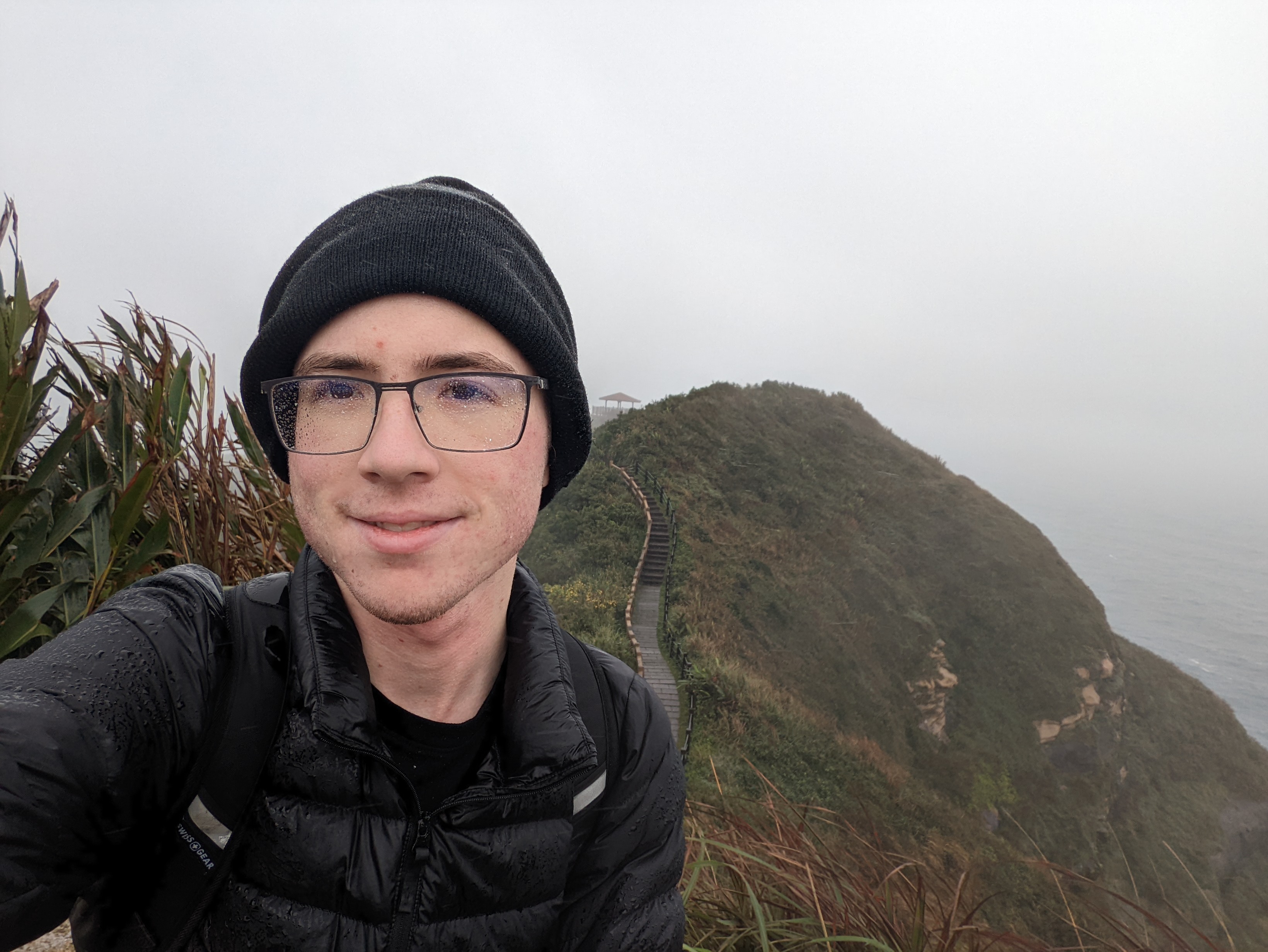 A selfie of me, Wesley Aptekar-Cassels, dsmiling at the camera in the rain and mist. There is a green hillside with a staircase on it visible in the background.