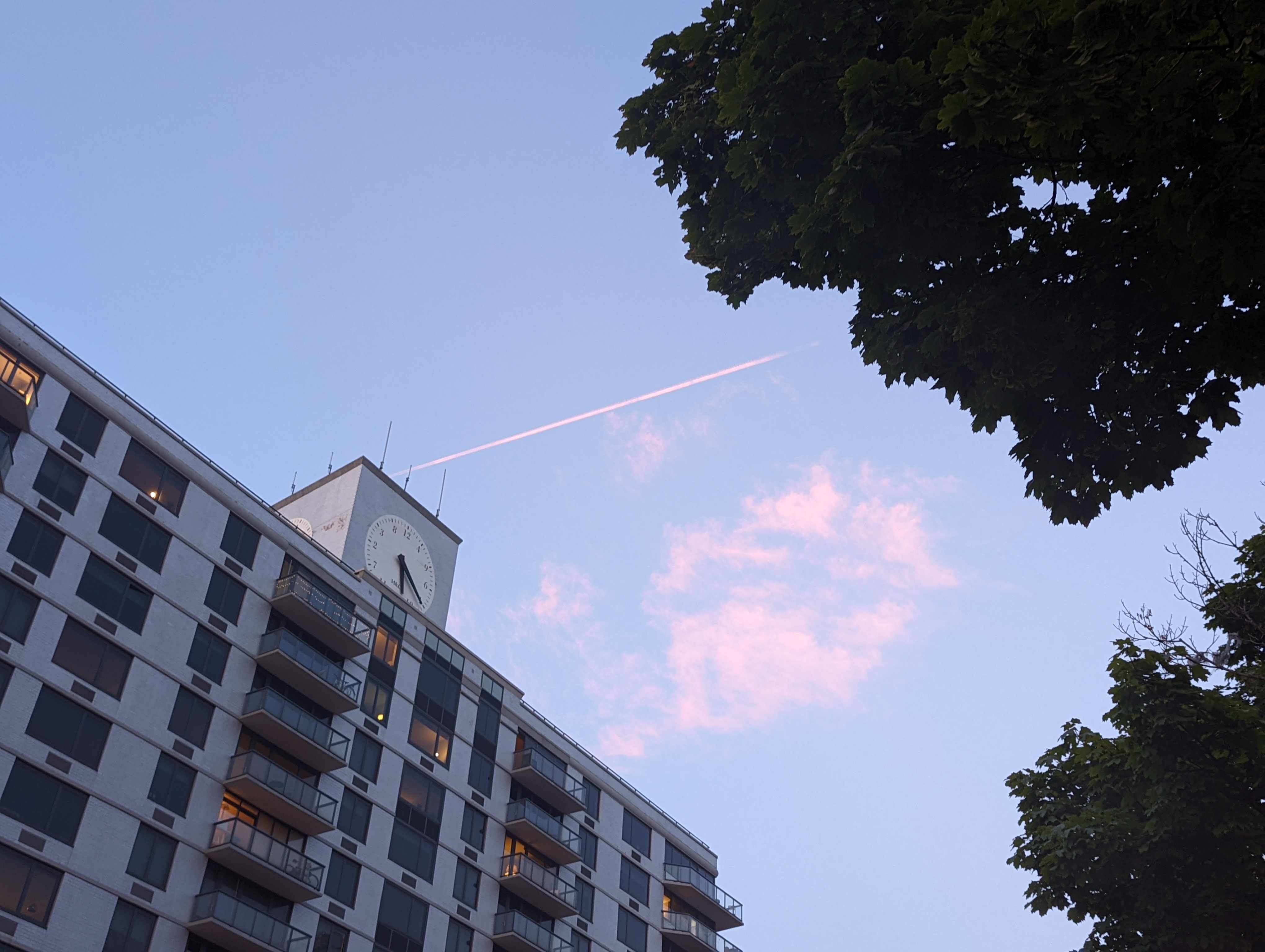 Photo of the sky, showing some pink clouds and a contrail, a building featuring a large clock, and foliage.