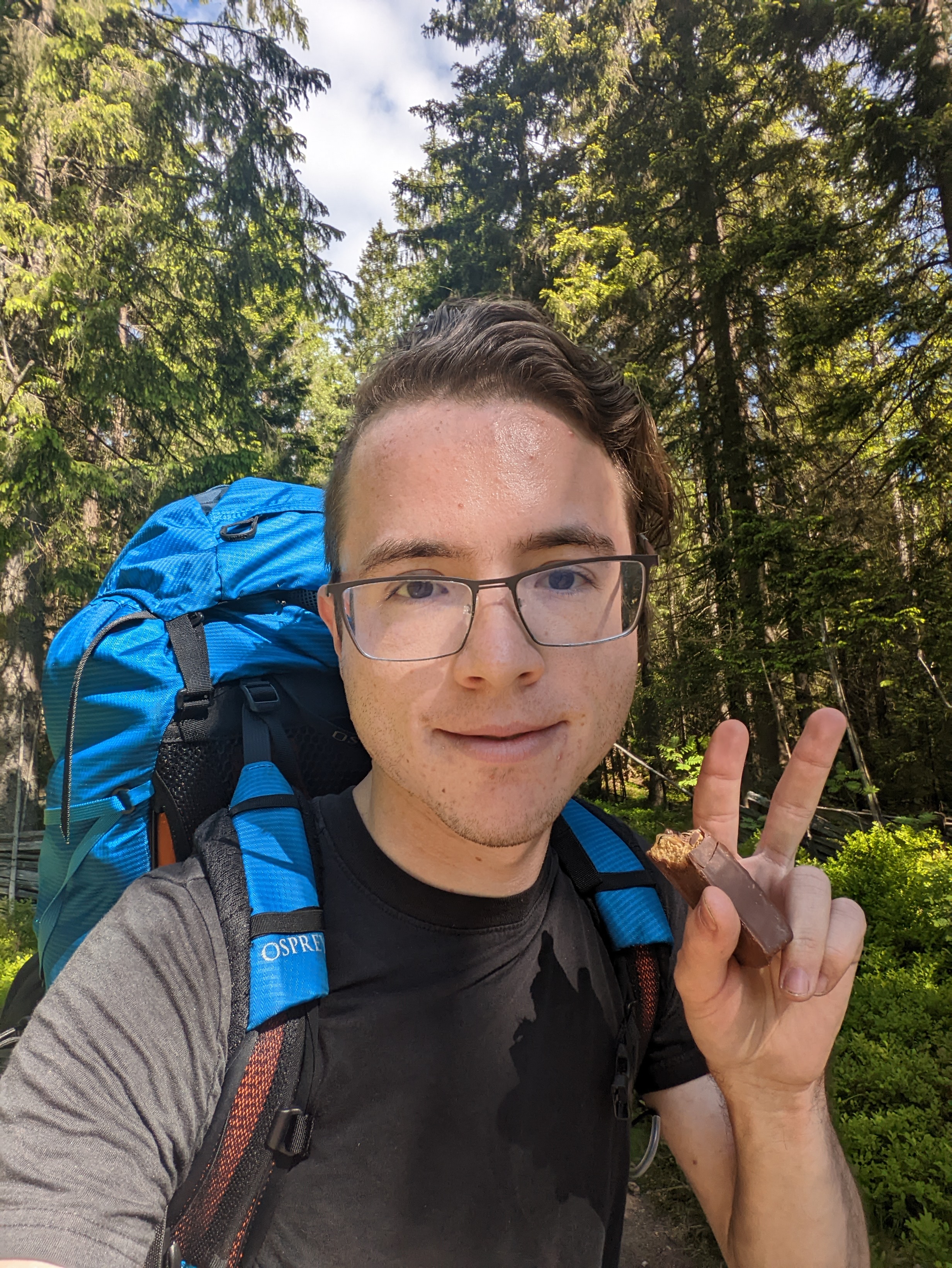 Photo of me, Wesley Aptekar-Cassels, wearing a backpacking backpack, making a peace sign, and holding a energy bar.
