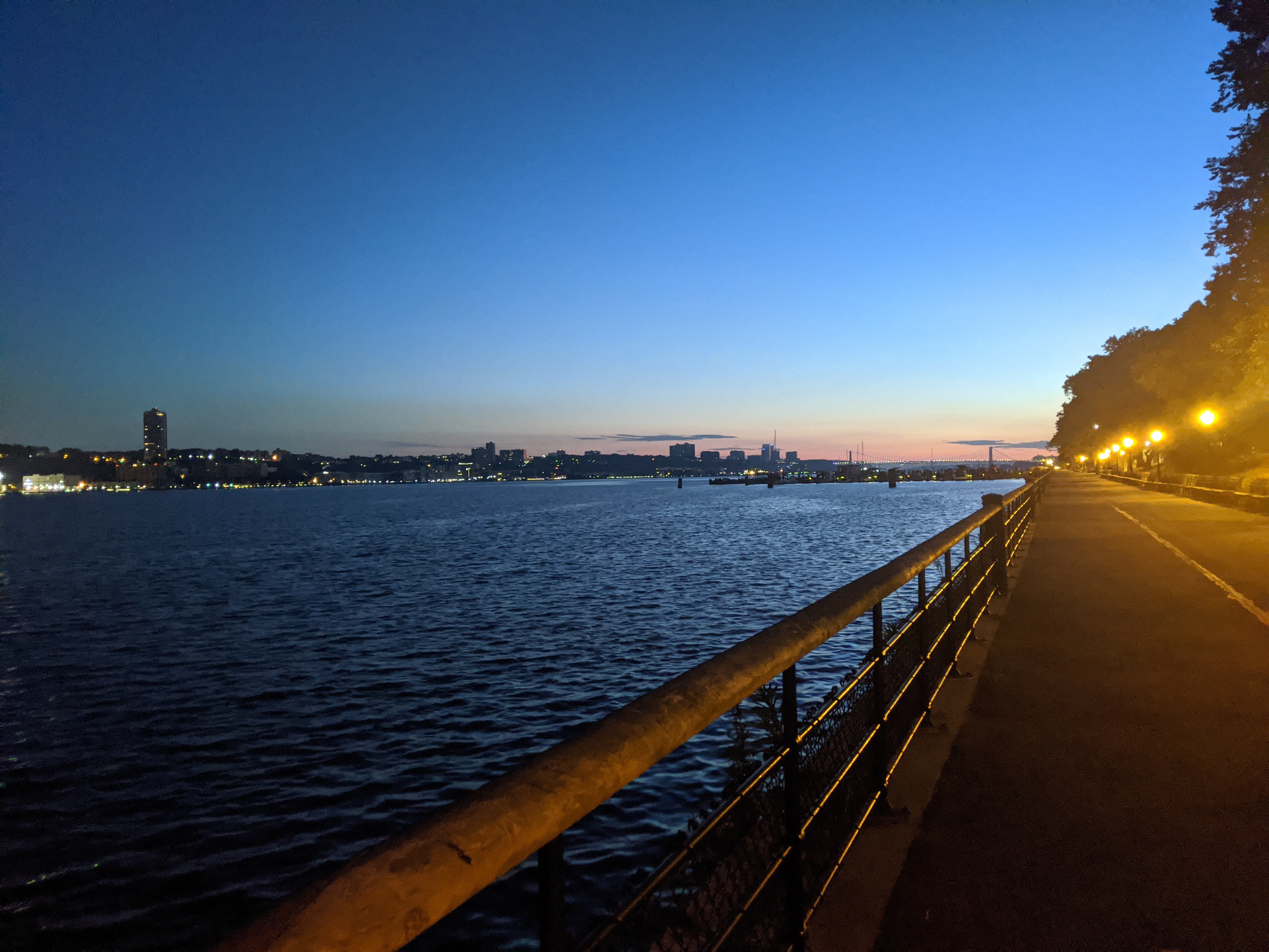 A photo of a walkway with a railing, next to a river. On the oppisite bank of the river is a city skyline. It's early in the morning, before the sun has fully risen, and streetlights provide much of the illumination in the photo.