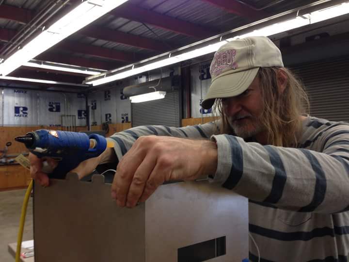 A photo ofSteve Inness, putting together a wooden or metal box, holding a industrial glue gun in one of his hands.